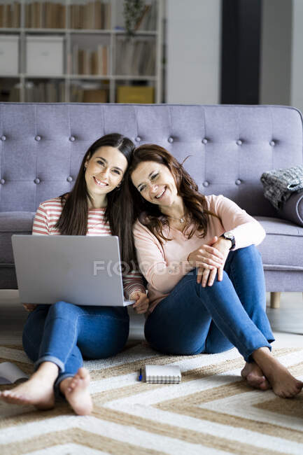 Smiling daughter with laptop sitting by mother on floor at home — Stock Photo