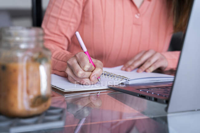 Mujer joven escribiendo en el diario mientras está sentada en la mesa de comedor en la oficina en casa - foto de stock