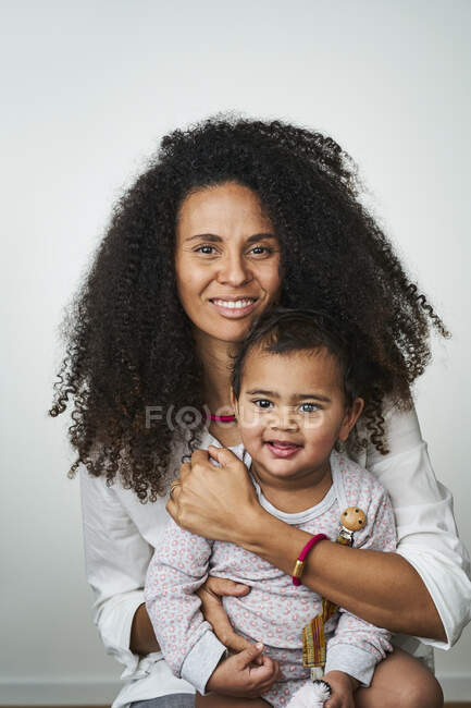 Sonriente mujer de pelo rizado llevando hija mientras se sienta sobre fondo gris - foto de stock