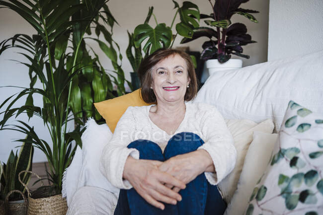 Femme souriante étreignant les genoux tout en étant assis sur le canapé à la maison — Photo de stock