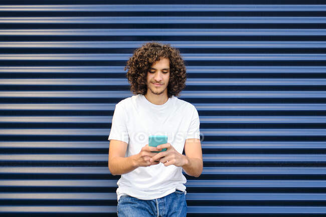 Молодий чоловік з кучерявим волоссям за допомогою смартфона спираючись на рифлене залізо. — стокове фото