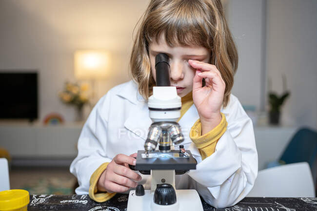 Chica en bata de laboratorio mirando a través del microscopio en casa - foto de stock