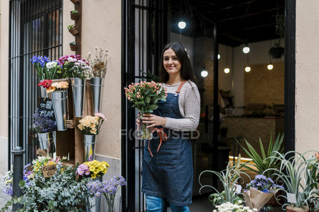 Young female florist holding flower vase while standing at shop — Stock Photo