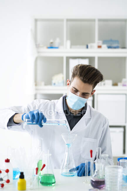 Hombre científico llenando matraz con medicina a través de embudo en laboratorio durante COVID-19 - foto de stock