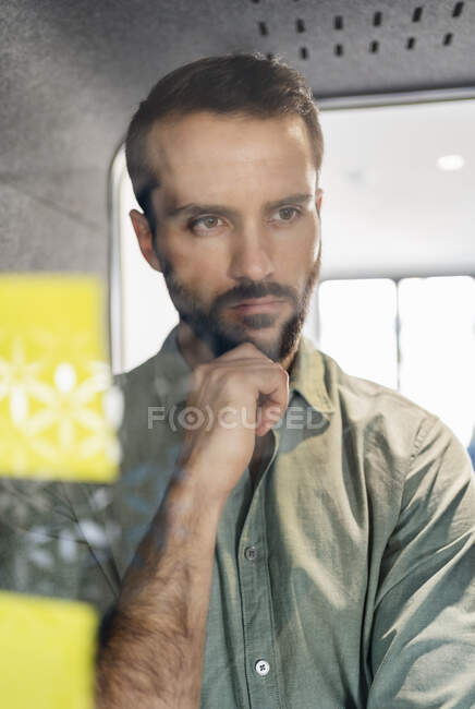 Thoughtful businessman looking at adhesive note on telephone booth glass at office — Stock Photo