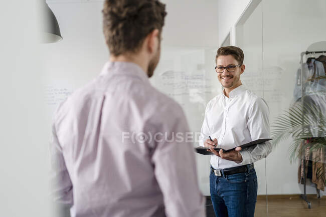 Улыбающийся бизнесмен с планшетом смотрит на коллегу-мужчину в офисе — стоковое фото