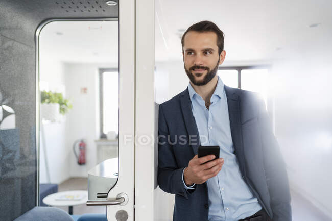 Young entrepreneur with mobile phone leaning on telephone booth at office — Stock Photo