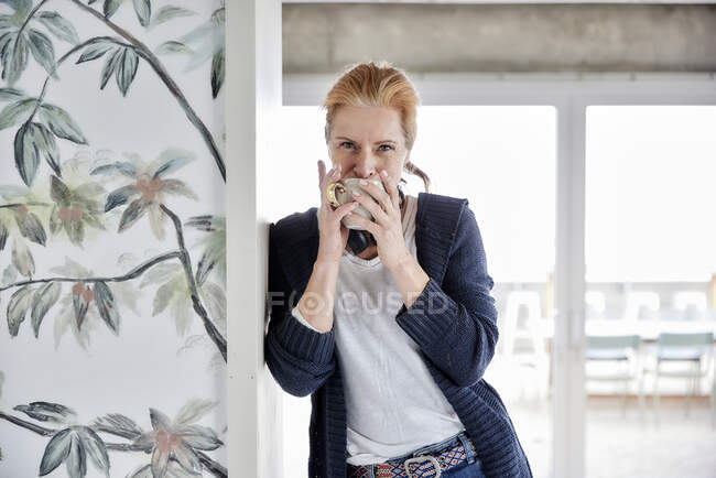 Frau trinkt Kaffee, während sie sich an Hauswand gegen Fenster lehnt — Stockfoto