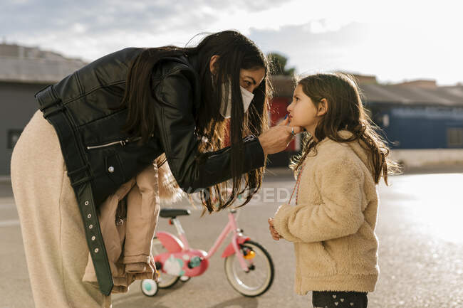 Madre indossa maschera protettiva viso pulizia viso di figlia mentre in piedi su strada — Foto stock