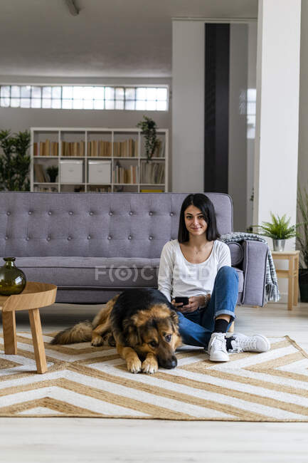 Усміхнена жінка сидить з собакою на килимі проти дивана у вітальні. — стокове фото