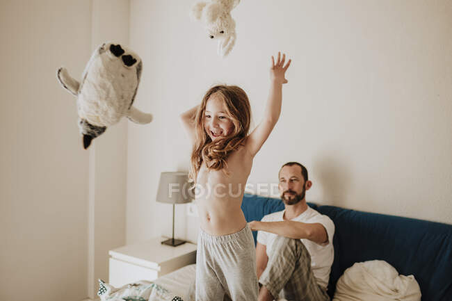 Весела дівчина кидає іграшки, а батько дивиться на ліжко. — стокове фото