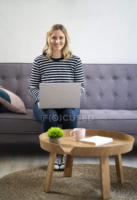Mujer rubia mediana adulta sentada con portátil en el sofá - foto de stock