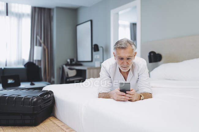 Hombre con anteojos acostado en la cama mientras usa el teléfono inteligente en la habitación de hotel — Stock Photo