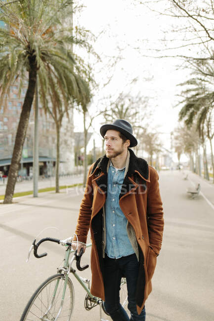 Hipster homem usando chapéu de pé com bicicleta na cidade enquanto olha para longe — Fotografia de Stock