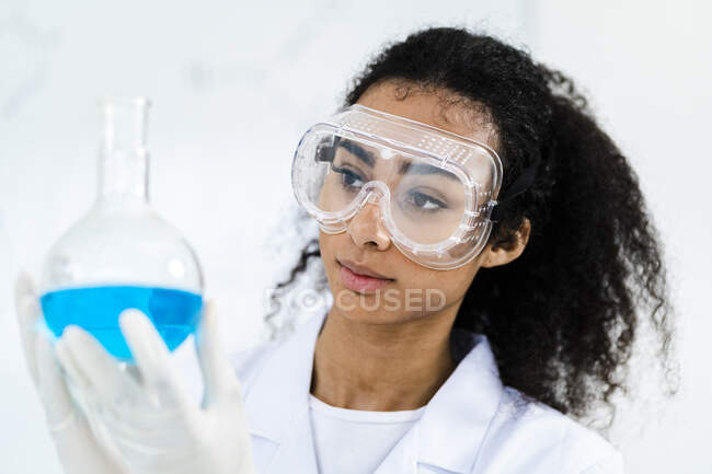 Joven investigadora en gafas protectoras examinando líquido en frasco en laboratorio - foto de stock