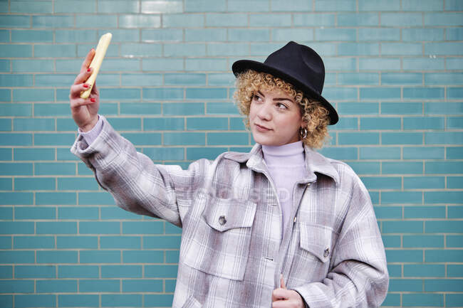Bella donna in cappello scattare selfie attraverso il telefono cellulare contro il muro blu — Foto stock