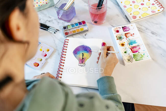 Mujer pintando bombilla con acuarelas en cuaderno espiral - foto de stock