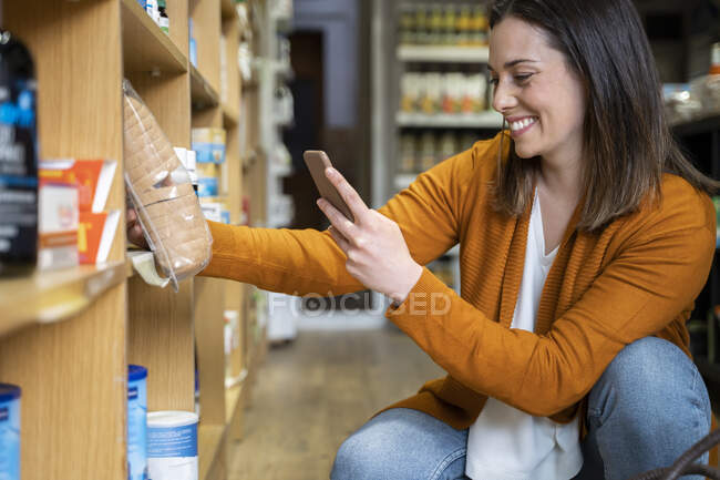 Happy young woman photographing package in grocery store — Stock Photo