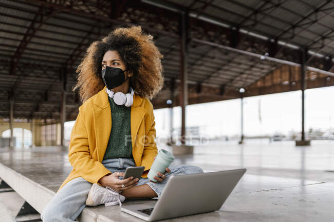 Afro-Frau mit Handy und Kaffeetasse schaut weg, während sie während der Pandemie mit Laptop auf der Sitzecke sitzt — Stockfoto