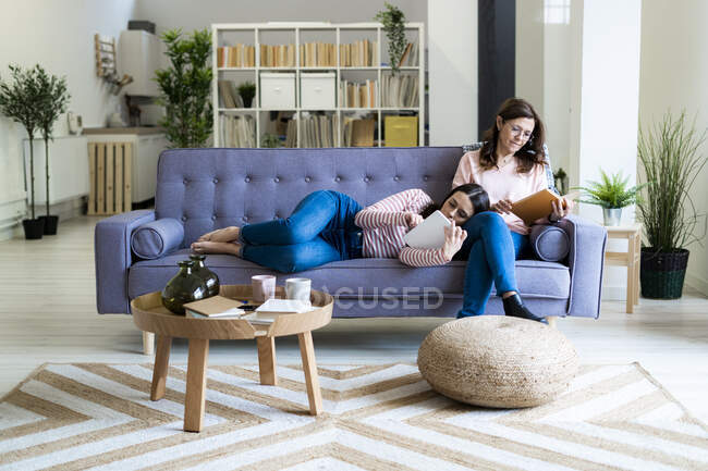 Madre lettura libro e figlia utilizzando tablet digitale mentre sdraiato sul divano a casa — Foto stock