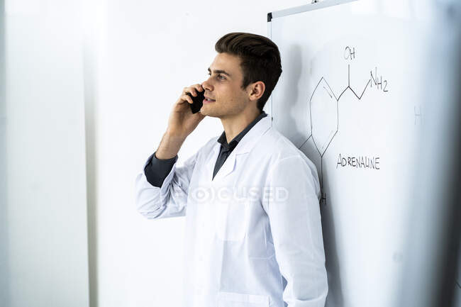 Jeune scientifique masculin parlant sur le téléphone intelligent tout en se tenant contre le tableau blanc en laboratoire — Photo de stock