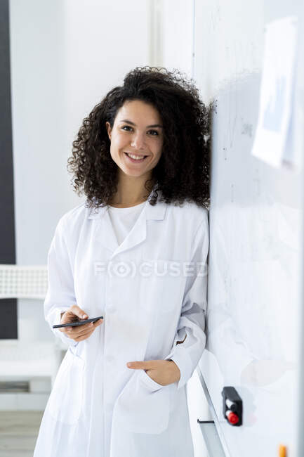 Cientista feminina sorridente com telefone inteligente inclinado por quadro branco no laboratório — Fotografia de Stock
