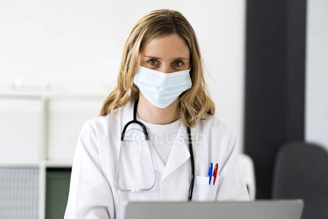 Mujer rubia trabajadora médica con laptop trabajando en clínica médica durante COVID-19 - foto de stock