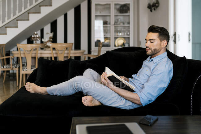 Mitte erwachsener Mann liest Buch, während er zu Hause auf dem Sofa sitzt — Stockfoto