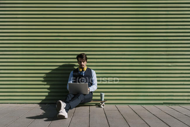 Empresario usando portátil mientras está sentado en el monopatín contra la pared verde - foto de stock