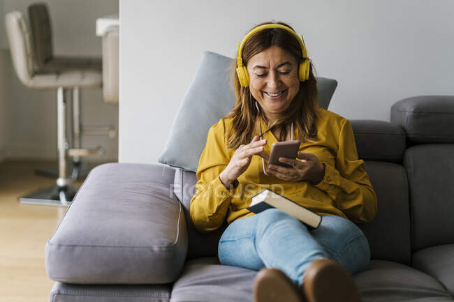 Smiling woman with headphones using smart phone while sitting on sofa in living room — Stock Photo