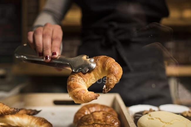 Jeune boulanger tenant croissant en servant des pinces à la boulangerie — Photo de stock
