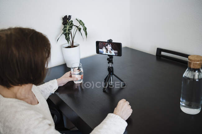 Woman holding glass of water while talking with daughter on mobile phone through video call — Stock Photo
