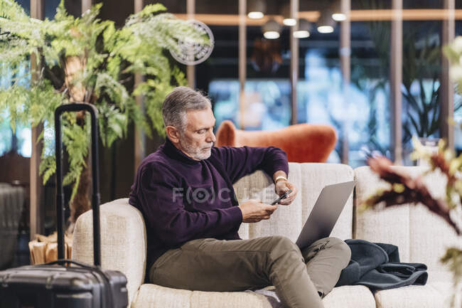 Зрілий підприємець за допомогою мобільного телефону сидить з ноутбуком на дивані в готелі. — стокове фото