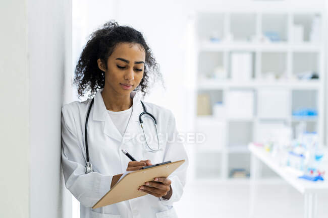 Jovem médica escrevendo na área de transferência enquanto se inclina na parede na clínica — Fotografia de Stock