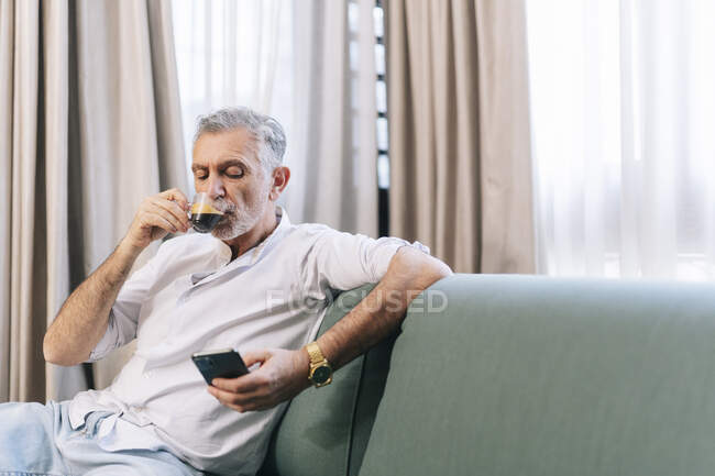 Reifer Mann beim Kaffee mit Smartphone, während er auf Sofa im Hotelzimmer sitzt — Stockfoto