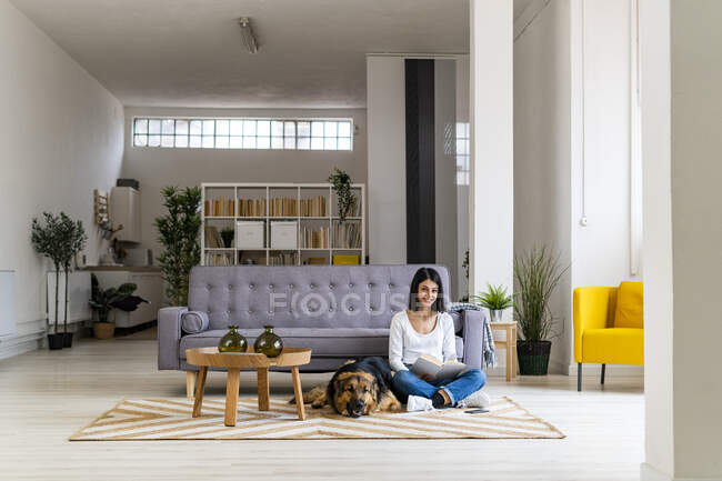 Femme souriante lisant le livre tout en étant assis avec chien de compagnie sur le tapis dans le salon — Photo de stock