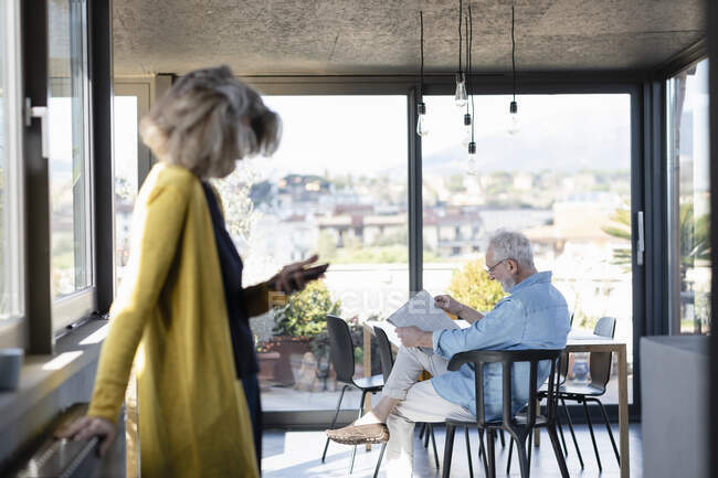 Donna in piedi alla finestra con l'uomo che legge il giornale seduto in background a casa — Foto stock