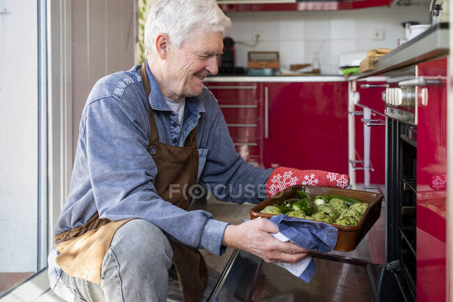 Sonriente hombre mayor poniendo bandeja de verduras en el horno - foto de stock