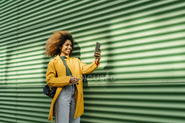 Улыбающаяся афро-женщина делает селфи через мобильный телефон, стоя у зеленой стены — стоковое фото