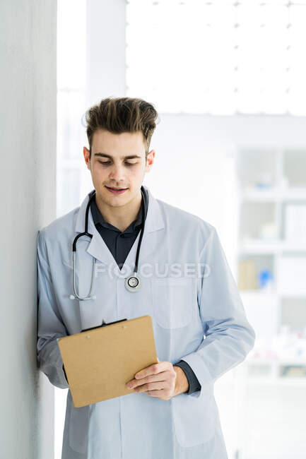 Male doctor writing notes while leaning on wall in hospital — Stock Photo