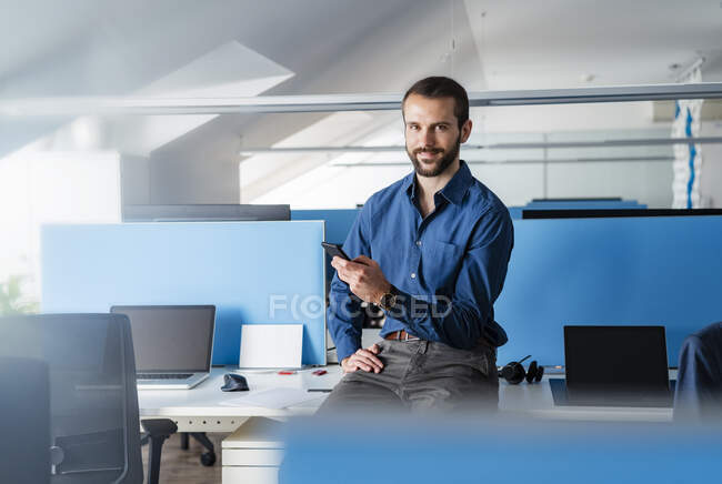 Uomo d'affari sorridente con cellulare seduto in ufficio — Foto stock
