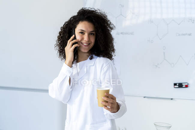 Médico sonriente con taza de café hablando por teléfono móvil en la clínica - foto de stock