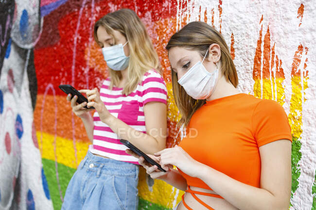 Amigas usando teléfono inteligente mientras se apoyan en la pared pintada durante COVID-19 - foto de stock