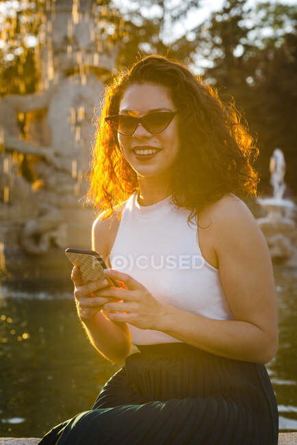 Smiling woman wearing sunglasses using smart phone while sitting against fountain during sunset — Stock Photo