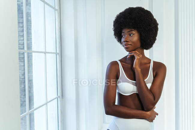 Modello afro femminile in lingerie con mano sul mento che guarda attraverso la finestra contro la parete bianca — Foto stock