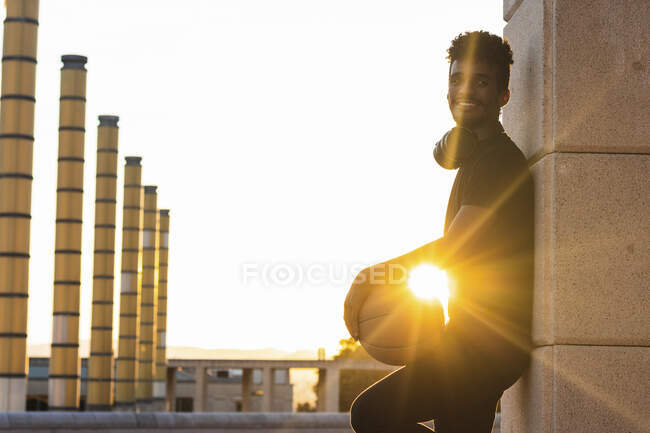Sun shining through smiling man holding basketball while standing by wall at sunset — Stock Photo