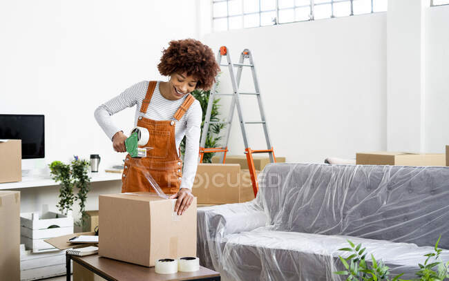 Afro woman packing box with adhesive tape while relocation in new home — Stock Photo