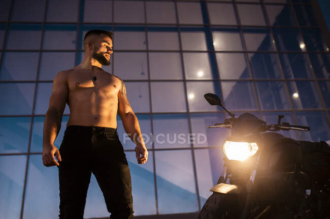 Shirtless handsome man staring motorcycle while standing against modern building — Stock Photo