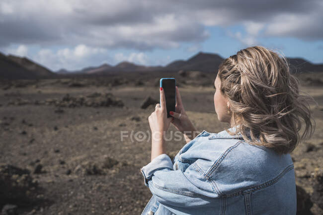 Молодая женщина делает фото через мобильный телефон, стоя на вулкане Эль-Куэрво, Лансароте, Испания — стоковое фото