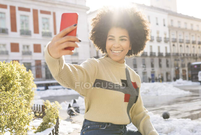 Afro woman smiling while taking selfie through mobile phone sitting in city — Stock Photo